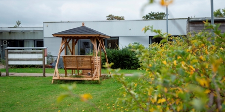 På billedet er et grønt område med en grå etplans bygning i baggrunden. Midt i billedet er der en dobbelt hængekøjesofa med tag over.