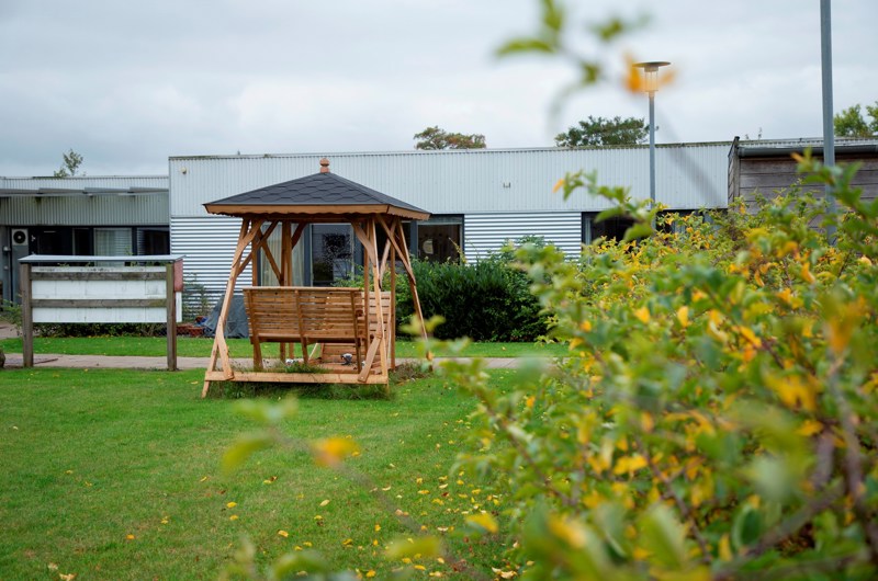 På billedet er et grønt område med en grå etplans bygning i baggrunden. Midt i billedet er der en dobbelt hængekøjesofa med tag over.