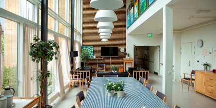 På billedet er der et opholdsrum med højt til loftet. I den ene halvdel er der 1. sal med udkig til opholdsrummet. Der er et langt spisebord i midten med 8 stole omkring og bagerst i lokalet er der en sofagruppe med 3 sofaer og TV. Til højre i lokalet er der gangsystem og til venstre er der vindue i hele lokalet fra gulv til loft.