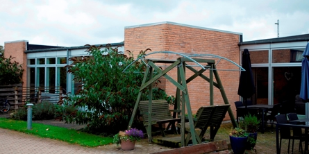 På billedet er et gult murstenshus, men mange vinduer i et plan, foran vinduerne er der små terrasser, med grønne planter omkring. Der er en dobbelt hængesofa og flere siddemiljøer på en flisebelagt terrasse og tre krukker med blomster foran huset.
