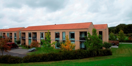 På billedet er der græsplæne i forgrunden og bagved ligger 3 store gulstens bygninger med store vinduespartier, foran bygningerne er lidt træer og plads til at parkere. Bag ved husene er der lidt skov til højre.