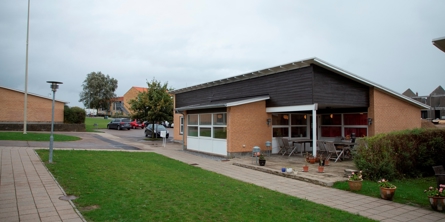 På billedet er der græs i forgrunden med fliser omkring. Til højre ligger et gult hus i et plan med overdækket terrasse integreret i huset. Bagved er der en parkeringsplads.