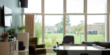 På billedet er et opholdsrum, med Airhocky  bagved er to lænestole og en sofa som står ved et stort vindue i hele bredden af rummet fra gulv til loft. Udsigten gennem vinduet er til et grønt område. I rummet er et TV og bogreoler.