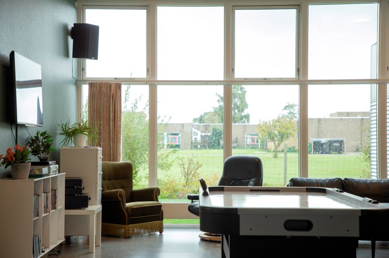 På billedet er et opholdsrum, med Airhocky  bagved er to lænestole og en sofa som står ved et stort vindue i hele bredden af rummet fra gulv til loft. Udsigten gennem vinduet er til et grønt område. I rummet er et TV og bogreoler.