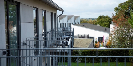 På billedet er en hvid bygning i forgrunden med store vinduespartier, med flere små altaner i stål. I baggrunden er flere hvide bygninger.