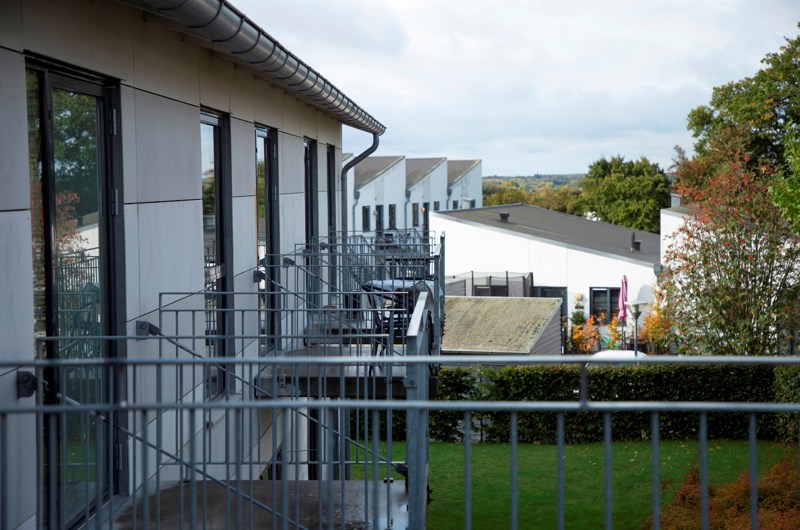 På billedet er en hvid bygning i forgrunden med store vinduespartier, med flere små altaner i stål. I baggrunden er flere hvide bygninger.