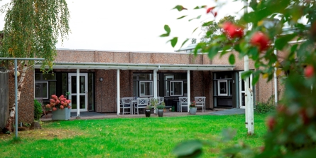 På billedet er en græsplæne i forgrunden og bagved et gråt hus med overdækket terrasse foran hvor der står hvide havemøbler. Foran terrassen er der en flagstang