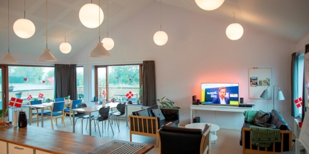 På billedet er et stort lyst lokale med loft til kip. I højre side er der sofagruppe med TV og i venstre er der to spiseborde med stole til 8 pers. Foran i lokalet er der et køkken og der er store vinduespartier i lokalet.