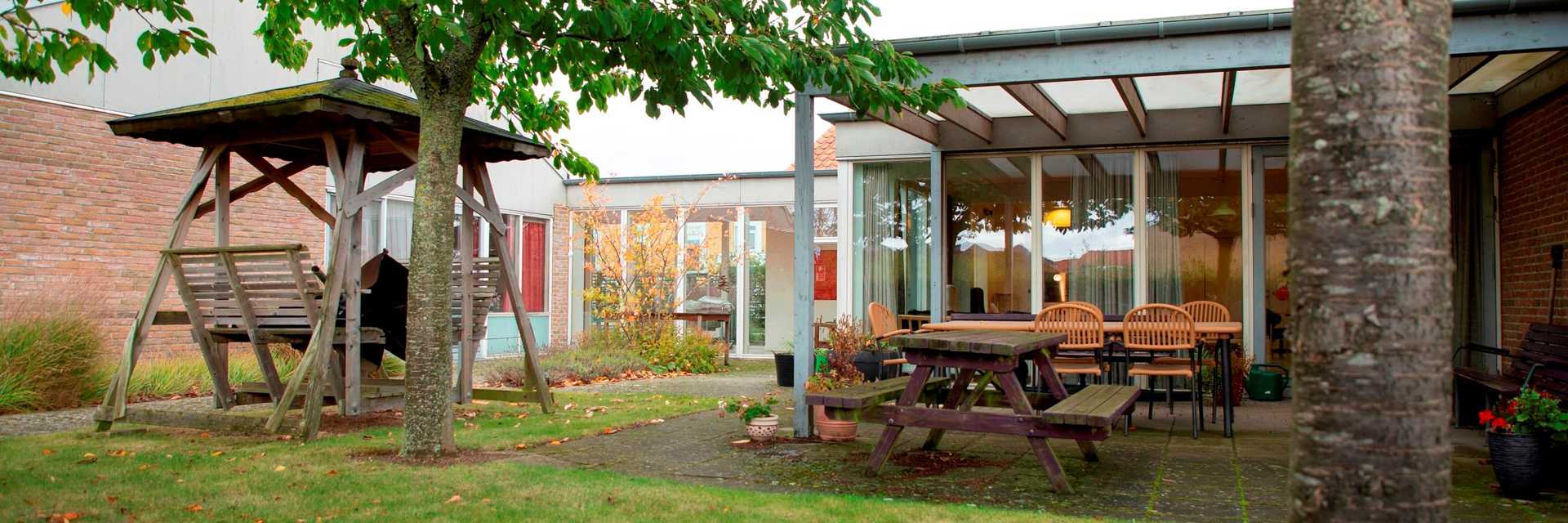 Billedet er taget udenfor ved Jasminvej 6-14. Der er græs i forgrunden og to træer der står forskudt, midt i billedet til højre står der en hængesofa i træ med et lille tag over og til højre er der siddegrupper med bænke borde og stole under en overdækket terrasse. I baggrunden er der en rød murstens bygning med store glaspartier