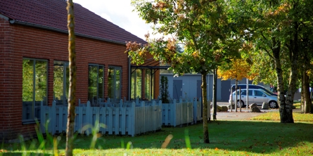 På billedet er der en græsplane med 3 små træer, til venstre er et rødt murstenshus med store vinduer i et plan. Foran vinduerne er små terrasser der er omkranset af lavt hvidt plankeværk