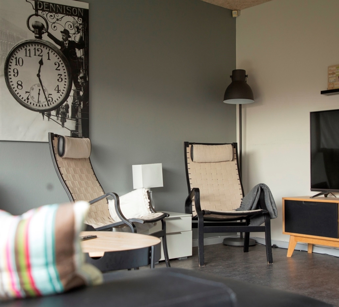 På billedet er et opholdsrum med sofa og to lænestole. Der er billeder på vægen og et TV. Den ene væg er malet grå og den anden hvid.